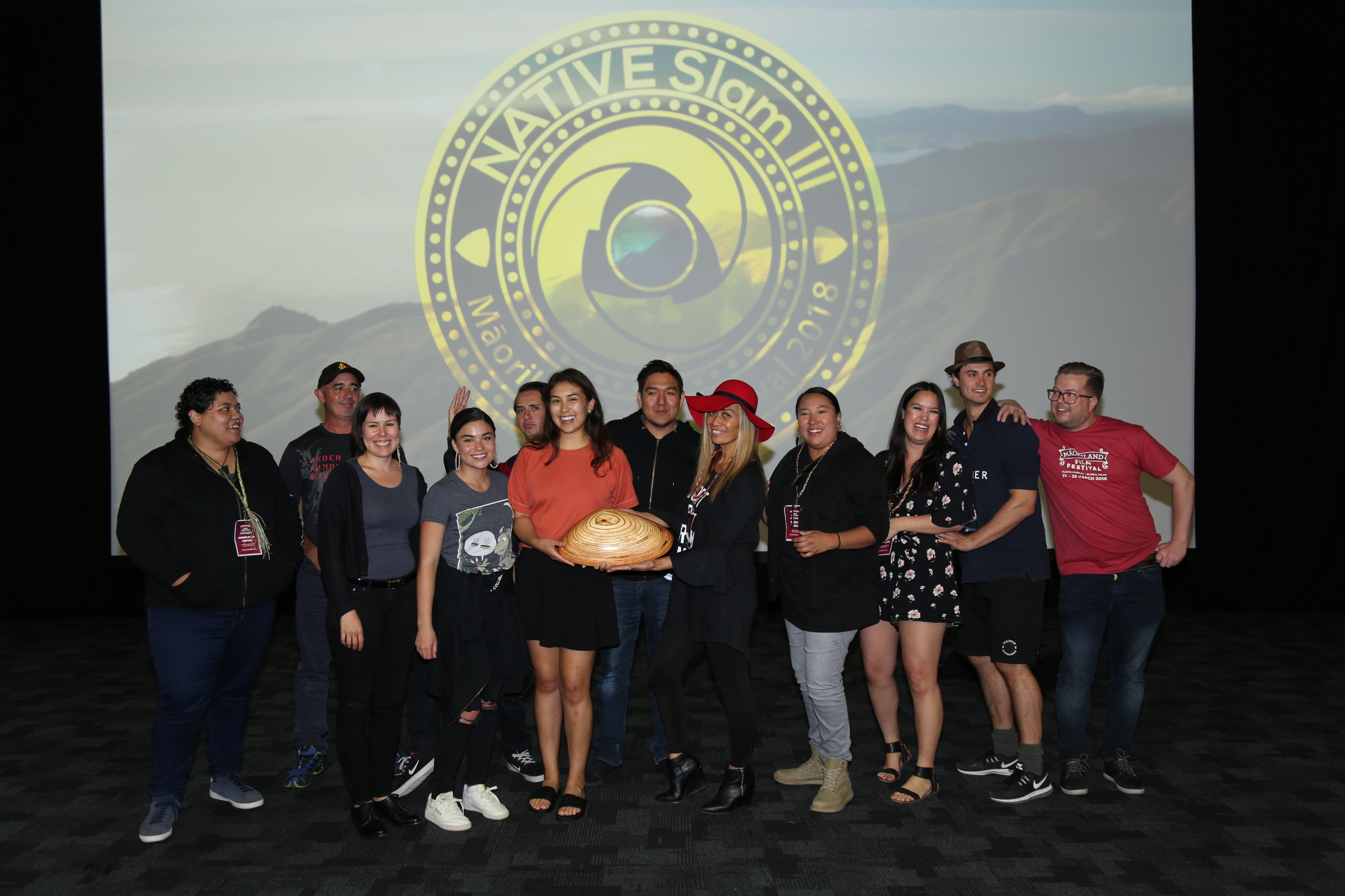 A group of filmmakers stand in front of a screen with the NATIVE Slam 3 logo behind them. In the centre a filmmaker holds a giant wooden pipi. 