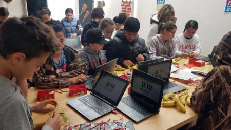 Rangatahi Māori crowd around a table with chromebooks and wires and bananas.