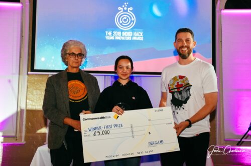 Young girl holds a giant cheque for $5,000 and is flanked on either side by adults. The cheque says "winner first prize" and in the background you can see "2019 Indigihack Young Innovators Awards"
