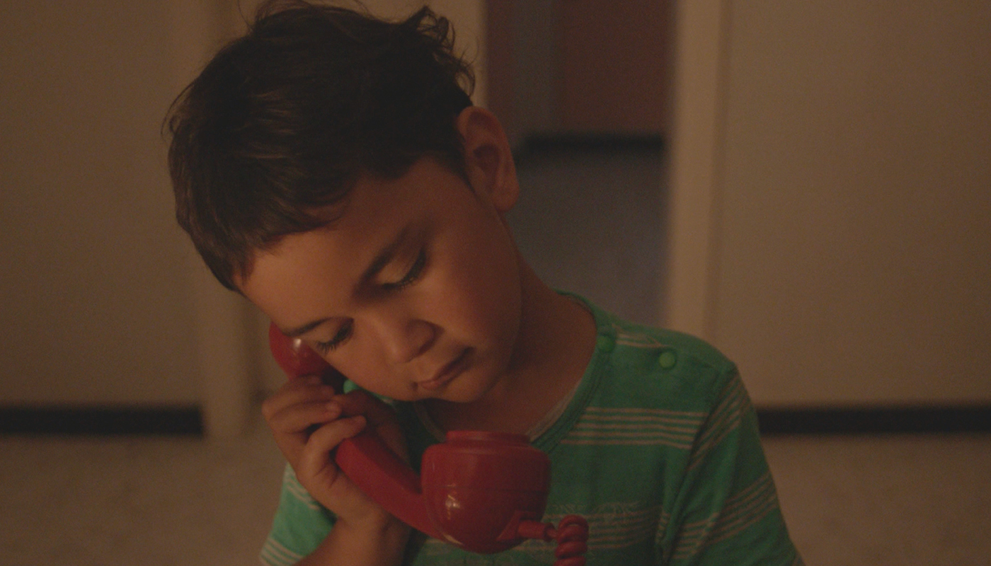 A small boy is pictured in a dim room on a red old school telephone. 