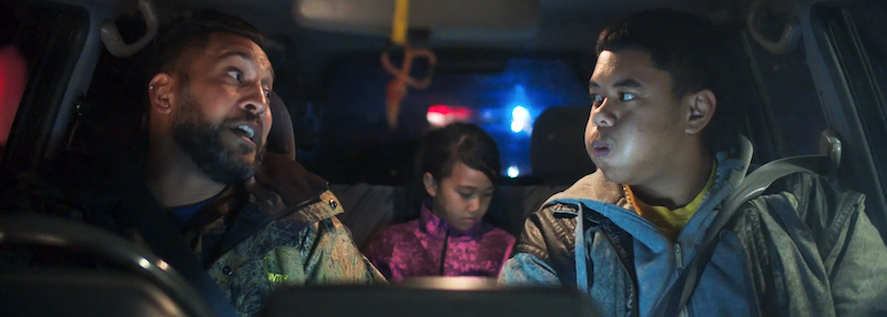 Image shows two Samoan men in the front seat of a car at night. A little girl sits in the back. The car is rear lit by what looks like a police car light.