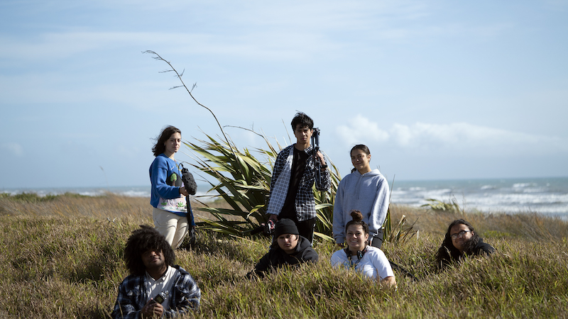 Image shows a group of teenagers standing broodily holding camera equipment on a wind swept grass dune with the beach in the background. 