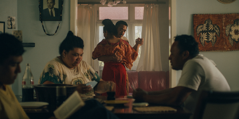 A Samoan couple slow dance in the background of a family sitting room. In the foreground other family members are reading. 