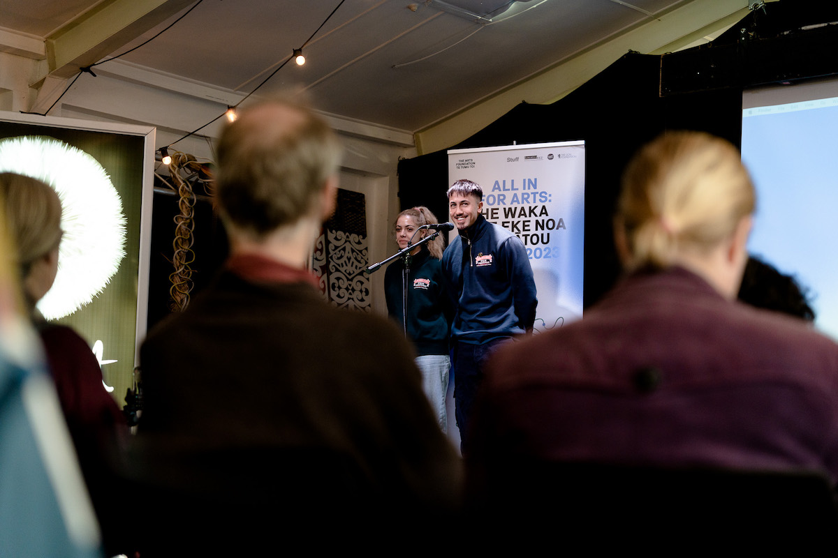 Matariki Black and Oriwa Hakaraia stand in front of a pull up banner labelled "All In For Arts" in front of an audience. They look cheery. 